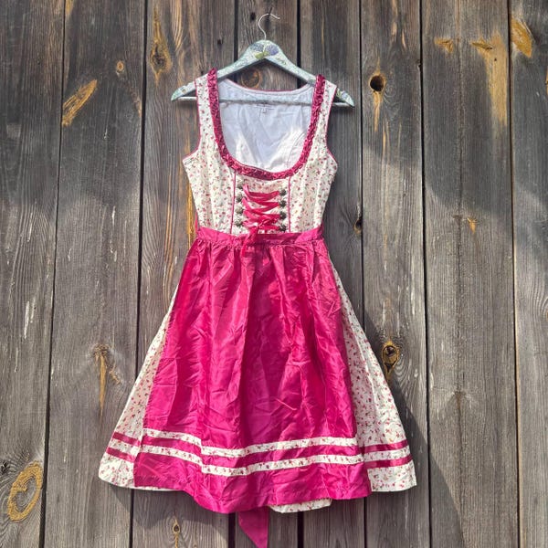 May include: A sleeveless dirndl dress featuring a white bodice with a small floral pattern, a deep pink satin skirt, and a matching pink lace-up front closure with decorative metal eyelets. The dress includes a white trim along the hemline.