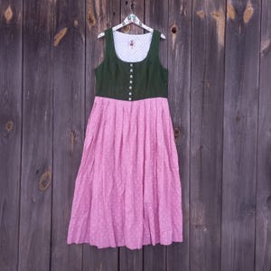 May include: A traditional dirndl dress, featuring a dark green bodice with white buttons and a pink skirt with a white polka dot pattern. The dress is sleeveless and hangs on a hanger against a wooden background.
