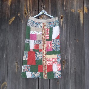 May include: A patchwork skirt with a variety of fabric patterns and colors, including floral, red, green, and white. The skirt is hanging on a decorative hanger against a wooden background. The skirt has a beige waistband and a vertical patterned strip down the center.