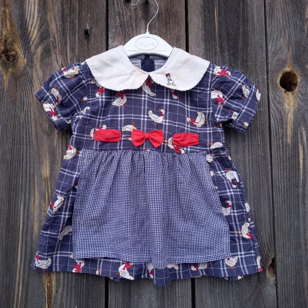 May include: A blue and white gingham dress with a white Peter Pan collar and a red bow. The dress features a pattern of white ducks.