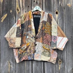 May include: A patchwork jacket with a variety of patterns and colors, including brown, beige, black, and orange. The jacket has a black lining and is fastened with three dark buttons. The jacket is hanging on a wooden background.