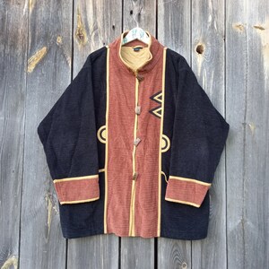 Könnte beinhalten: Eine schwarze und rostfarbene Jacke mit einem Stehkragen und dekorativen Knöpfen. Die Jacke hat ein geometrisches Muster auf der Vorderseite und einen braunen Besatz an den Ärmelbündchen und am Kragen.