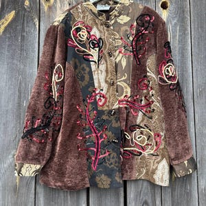 May include: Brown velvet jacket with floral embroidery and sequin accents. The jacket features a mix of patterned fabrics and a button-up front. The sleeves and collar have a patterned trim. The jacket is hanging on a wooden background.