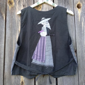 May include: A black vest with white polka dots and a decorative appliqué of a woman in a hat and dress. The vest has a striped pocket and a tie closure at the waist. The woman's dress has purple and grey accents.