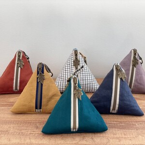 May include: A collection of triangular fabric pouches in various colors, including rust, mustard, teal, navy, and lavender. Each pouch features a contrasting zipper with a leaf-shaped pull and a vertical stripe detail.