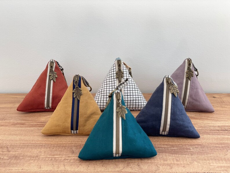May include: A collection of triangular fabric pouches in various colors, including rust, mustard, teal, navy, and lavender. Each pouch features a contrasting zipper with a leaf-shaped pull and a vertical stripe detail.