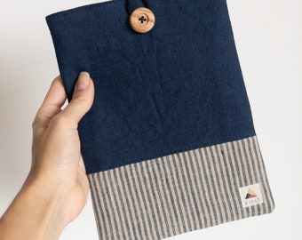 Pode incluir: Uma capa de tecido azul marinho e listrado com fecho de botão de madeira. A parte superior é azul marinho liso, enquanto a parte inferior apresenta listras verticais cinza e brancas. Uma pequena etiqueta com o texto "VIVIT" é visível.