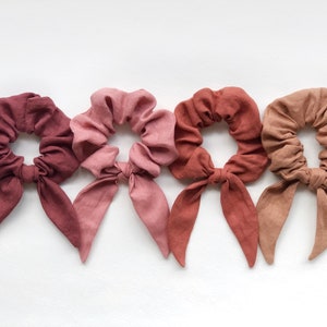 May include: Four scrunchies in shades of red and pink. Each scrunchie has a matching fabric bow. The scrunchies are made of a soft, textured material and are arranged in a row against a white background. These hair accessories are perfect for adding a touch of color to any hairstyle.