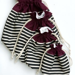 May include: Four drawstring bags in varying sizes. The bags have a burgundy top and a black and white striped lower section. Each bag has a white drawstring. The bags are arranged on a white background.