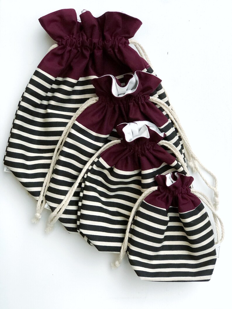 May include: Four drawstring bags in varying sizes. The bags have a burgundy top and a black and white striped lower section. Each bag has a white drawstring. The bags are arranged on a white background.
