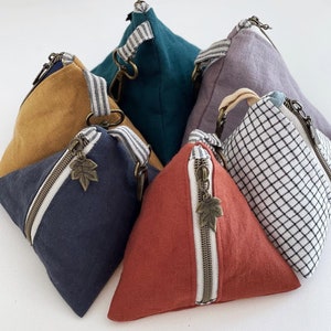 May include: A collection of triangular pouches in various colors, including navy, mustard, teal, and red. Each pouch features a diagonal zipper with a leaf charm, a striped fabric handle, and a metal zipper. The pouches are made of fabric.
