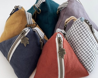 May include: A selection of triangular pouches in shades like navy, mustard, teal, and red. Each pouch has a diagonal zip with a leaf charm, a striped fabric handle, and a metal zip. The pouches are made of fabric.