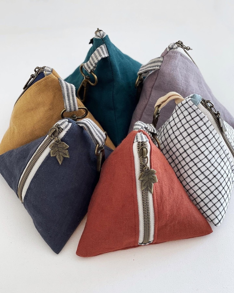 May include: A collection of triangular pouches in various colors, including navy, mustard, teal, and red. Each pouch features a diagonal zipper with a leaf charm, a striped fabric handle, and a metal zipper. The pouches are made of fabric.