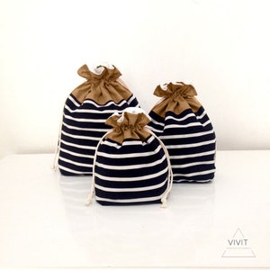 May include: Three drawstring bags in varying sizes. The bags feature a navy blue and white striped pattern on the lower portion, with a tan-colored top and white interior. Each bag has a white drawstring closure.