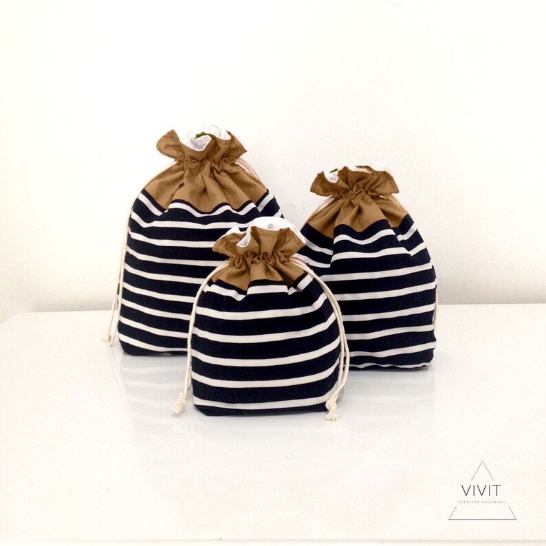 May include: Three drawstring bags in varying sizes. The bags feature a navy blue and white striped pattern on the lower portion, with a tan-colored top and white interior. Each bag has a white drawstring closure.