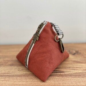 May include: A small, triangular rust-colored pouch with a bronze zipper and leaf-shaped pull. The zipper runs along one side, and a striped fabric loop with a bronze clasp is attached. The pouch is made of a textured fabric.