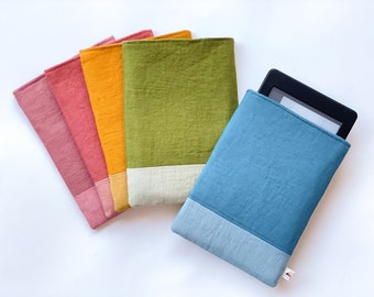 May include: A collection of colorful fabric e-reader cases. The cases are in shades of pink, orange, green, and blue, each with a contrasting bottom panel. One case holds a black e-reader device. The cases are made of a textured fabric.