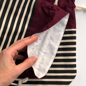 May include: A drawstring bag with a striped pattern in black and cream, and a solid burgundy top. The interior is white with stitched details. The bag is made of fabric and has a cream-colored drawstring.