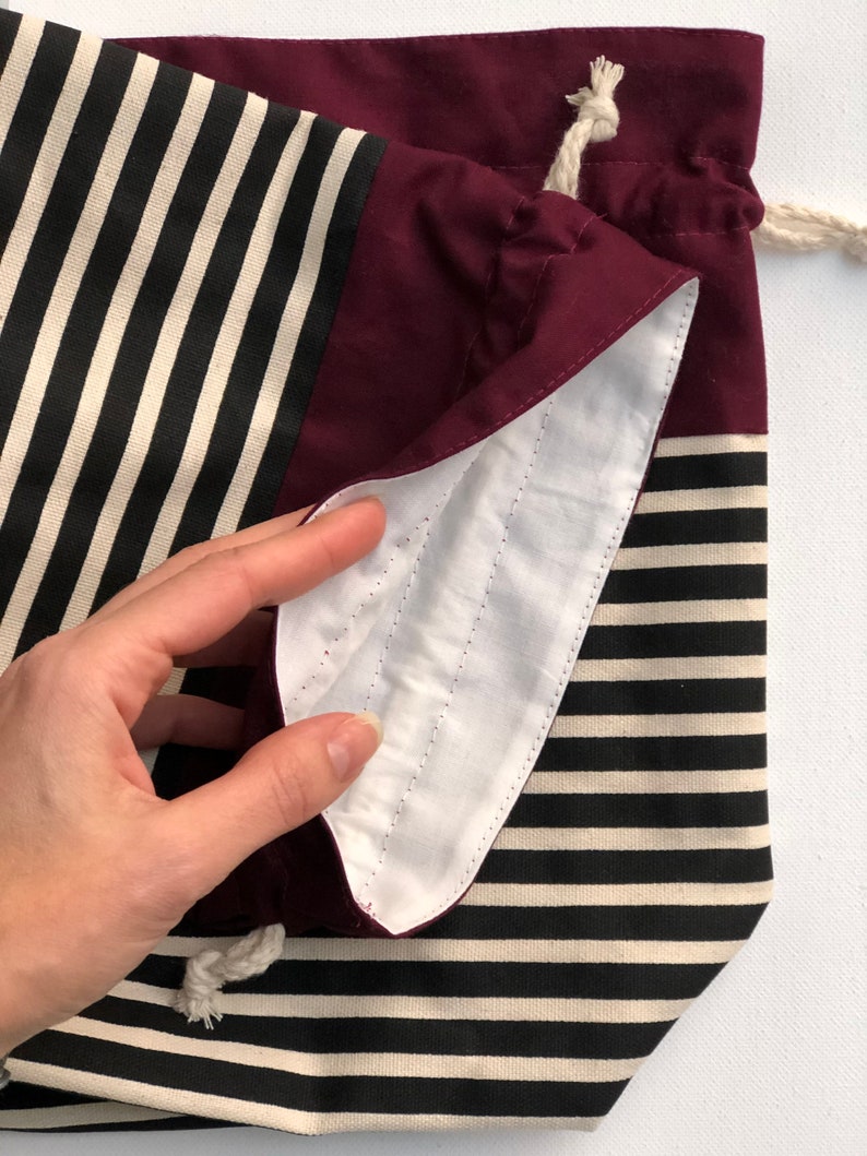 May include: A drawstring bag with a striped pattern in black and cream, and a solid burgundy top. The interior is white with stitched details. The bag is made of fabric and has a cream-colored drawstring.