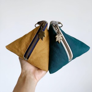 May include: Two triangular pouches, one mustard yellow and one teal, are displayed. Each pouch has a zipper with a leaf-shaped charm and a small loop. The mustard pouch has a navy blue stripe, while the teal pouch has a cream stripe.