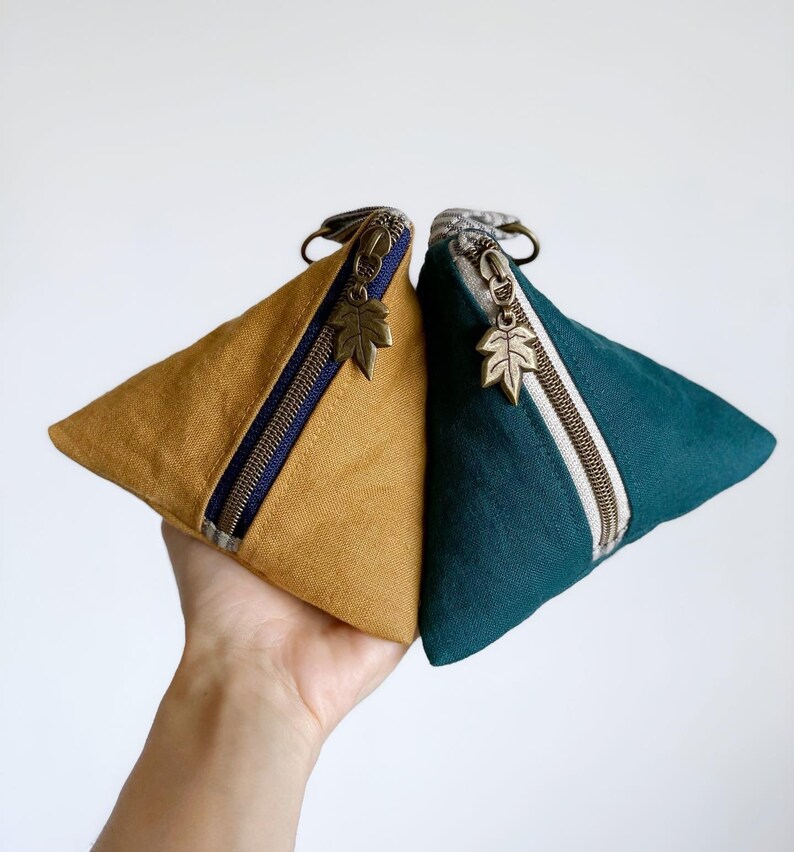 May include: Two triangular pouches, one mustard yellow and one teal, are displayed. Each pouch has a zipper with a leaf-shaped charm and a small loop. The mustard pouch has a navy blue stripe, while the teal pouch has a cream stripe.
