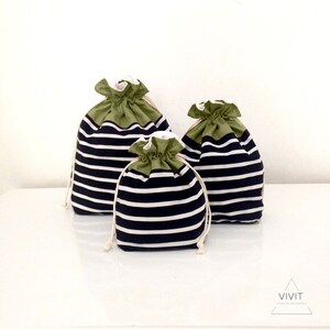 May include: Three drawstring bags of varying sizes. The bags have a navy blue and white striped pattern on the body and olive green tops. The drawstring is a natural off-white color. The bags are sitting on a white surface.