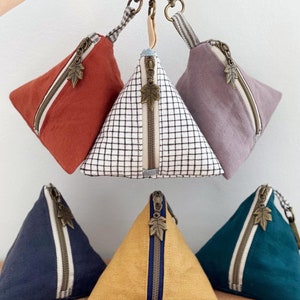 May include: A collection of pyramid-shaped coin purses in various colors, including rust, navy, and mustard yellow. Each purse features a diagonal zipper with a leaf-shaped pull and a striped accent. The purses are attached to a keychain.