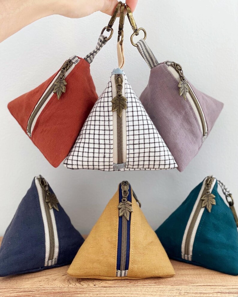 May include: A collection of pyramid-shaped coin purses in various colors, including rust, navy, and mustard yellow. Each purse features a diagonal zipper with a leaf-shaped pull and a striped accent. The purses are attached to a keychain.