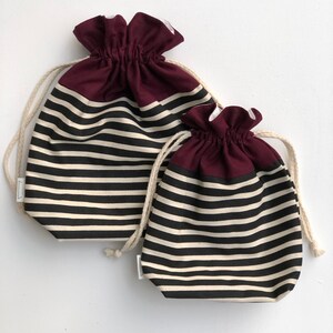 May include: Two drawstring bags with a striped pattern in black and off-white. The top section of each bag is a solid burgundy color. The bags have a natural-colored drawstring closure.