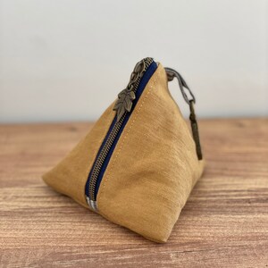 May include: A tan, triangular-shaped pouch with a dark blue zipper and a leaf-shaped zipper pull. The pouch is made of a textured fabric and sits on a wooden surface. The zipper is accented with a blue and white striped fabric.