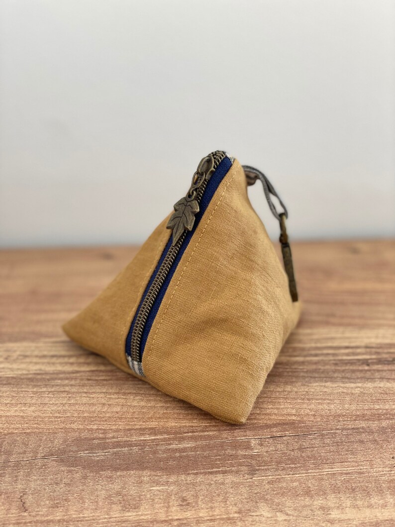 May include: A tan, triangular-shaped pouch with a dark blue zipper and a leaf-shaped zipper pull. The pouch is made of a textured fabric and sits on a wooden surface. The zipper is accented with a blue and white striped fabric.