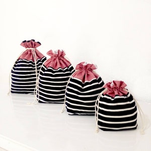 May include: Four drawstring bags in varying sizes. The bags have a navy blue and white striped pattern on the body and a dusty rose colored top. Each bag has a white drawstring closure.