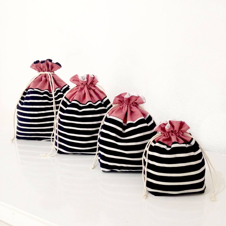 May include: Four drawstring bags in varying sizes. The bags have a navy blue and white striped pattern on the body and a dusty rose colored top. Each bag has a white drawstring closure.