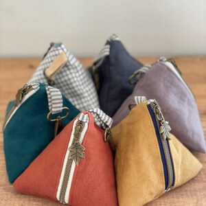 May include: A collection of triangular fabric pouches in various colors, including teal, rust, and mustard yellow. Each pouch features a metal zipper with a leaf-shaped pull and a striped accent. The pouches are arranged on a wooden surface.