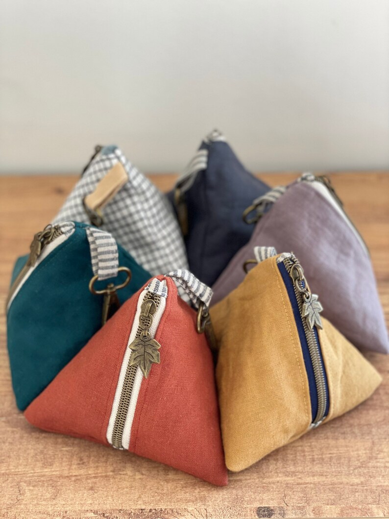 May include: A collection of triangular fabric pouches in various colors, including teal, rust, and mustard yellow. Each pouch features a metal zipper with a leaf-shaped pull and a striped accent. The pouches are arranged on a wooden surface.