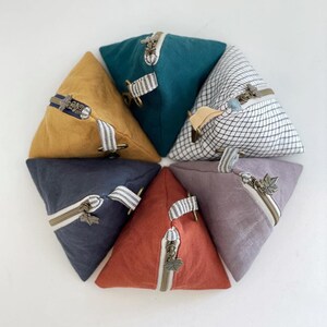 May include: A collection of triangular fabric pouches in various colors, including mustard yellow, teal, and rust. Each pouch features a zipper closure with a decorative pull and a fabric loop. The pouches are arranged in a circular pattern.
