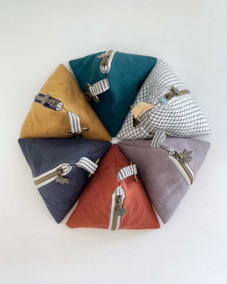 May include: A collection of triangular fabric pouches in various colors, including mustard yellow, teal, and rust. Each pouch features a zipper closure with a decorative pull and a fabric loop. The pouches are arranged in a circular pattern.