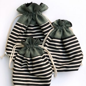May include: Three drawstring bags in varying sizes. The bags have a dark green top and a black and white striped pattern on the body. Each bag has a natural-colored drawstring closure.