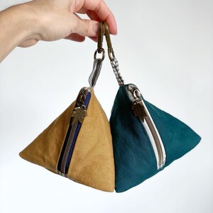 May include: Two triangular pouches with brass-colored zippers and leaf-shaped zipper pulls. One pouch is mustard yellow, the other is teal. Each has a small fabric loop attached to a brass-colored clasp.
