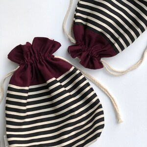 May include: Two drawstring bags with a black and cream horizontal striped pattern. The bottom of each bag is a solid burgundy color. The bags have a gathered top and a cream-colored drawstring. The bags are on a white background.
