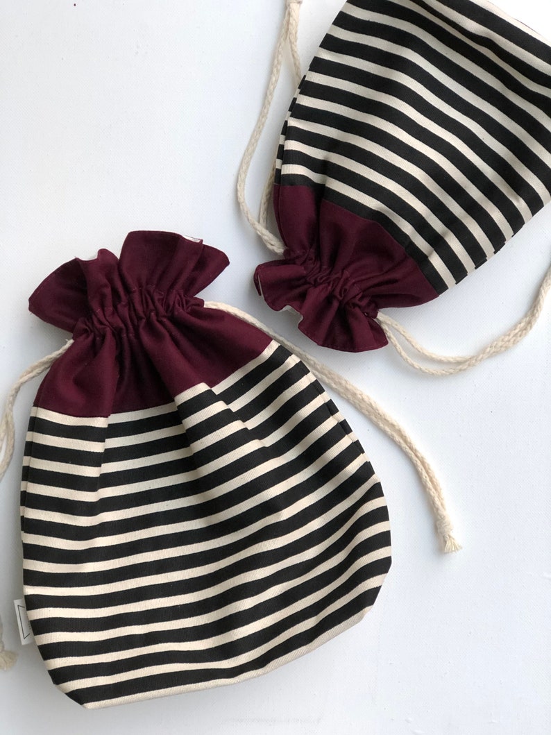 May include: Two drawstring bags with a black and cream horizontal striped pattern. The bottom of each bag is a solid burgundy color. The bags have a gathered top and a cream-colored drawstring. The bags are on a white background.