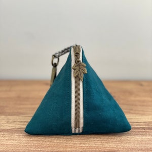 May include: Teal triangular pouch with a vertical white stripe and a bronze zipper. The zipper pull is a leaf charm. The pouch has a small loop and a braided strap attached to the side. The pouch is made of a textured fabric.