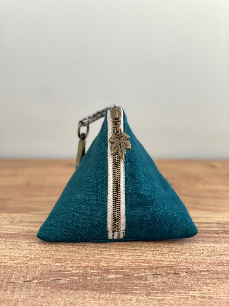 May include: Teal triangular pouch with a vertical white stripe and a bronze zipper. The zipper pull is a leaf charm. The pouch has a small loop and a braided strap attached to the side. The pouch is made of a textured fabric.