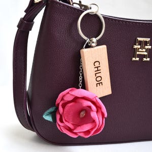 Peut inclure: Un sac à main prune avec un porte-clés argenté et une étiquette en bois avec le nom "CHLOE". Une fleur en tissu rose avec une feuille verte est suspendue au porte-clés. Le sac a un emblème doré.