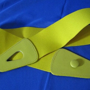 May include: A bright yellow elastic belt with lime green leather accents. The belt features a buckle with a unique cut-out design and a matching button closure. The belt is shown against a blue background.