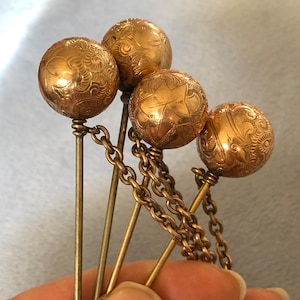 May include: Four antique gold-toned hat pins with decorative ball heads. The ball heads feature intricate embossed patterns and are connected by a delicate chain. The pins are held together, showcasing their vintage design.