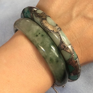May include: Two jade bangles. One is a solid, glossy green. The other is light green with silver filigree and turquoise accents. The bangles are worn together on a wrist, showcasing their contrasting designs and textures.