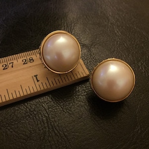 Puede incluir: Un par de pendientes de clip de tono dorado con grandes piedras redondas de imitación de perla blanca.