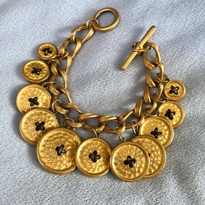 Puede incluir: Un brazalete de cadena dorada con cierre de palanca y múltiples dijes de botón dorado. Cada dije de botón tiene un centro negro.