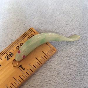 May include: A jade green fish pendant with red gemstone eyes and a gold bail. The pendant is approximately 1 inch long.